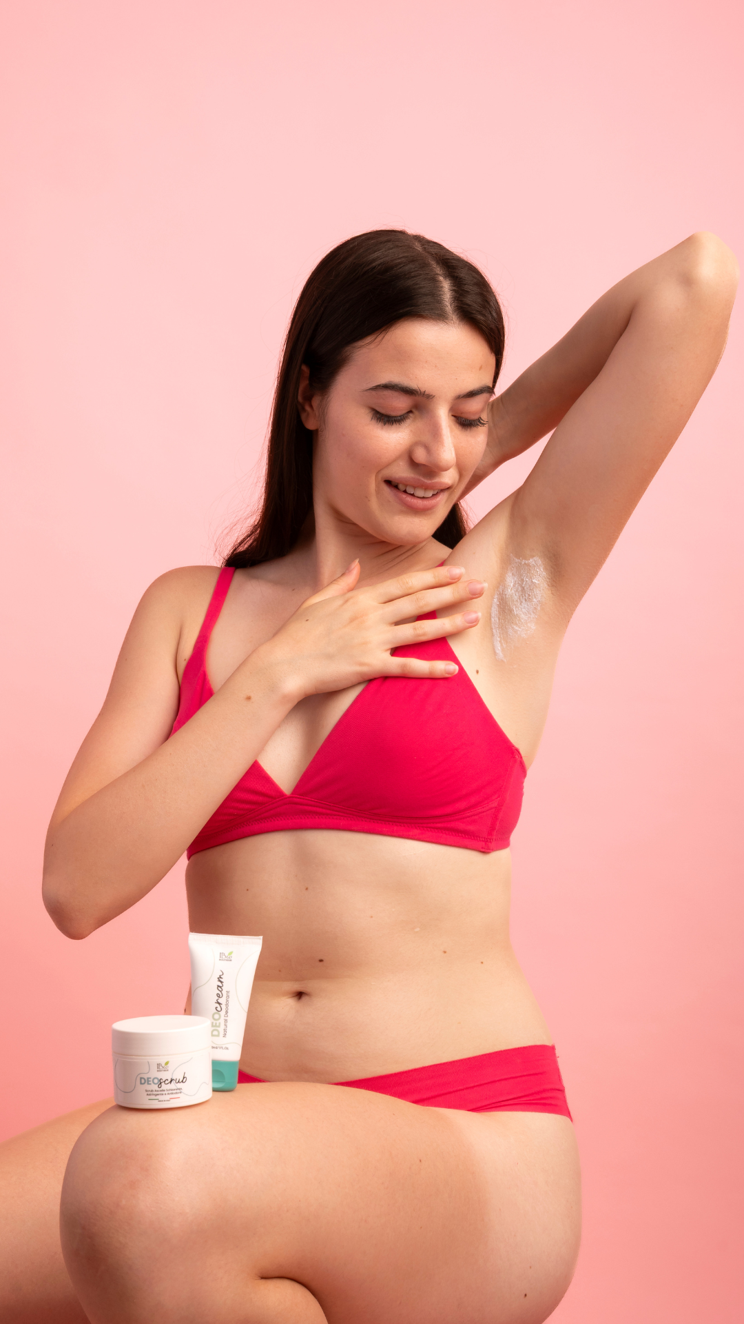 Ragazza castana in posizione rannicchiata mette dolcemente una crema sull'ascella per combattere il cattivo odore dovuto al sudore acido